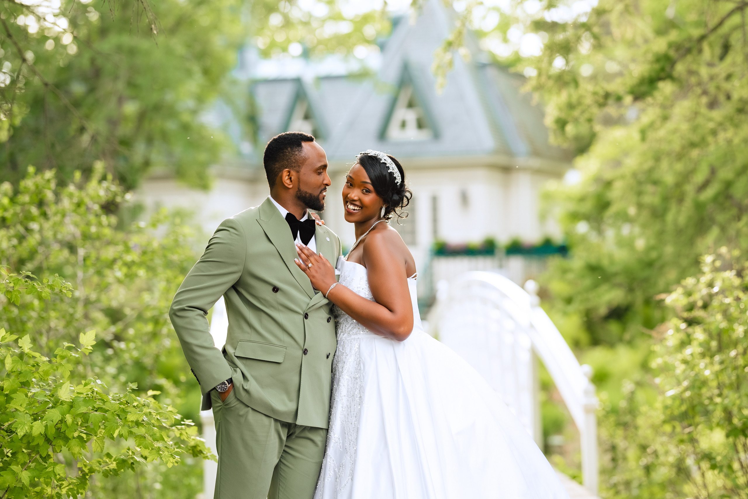 A groom kisses the bride near her temple as she smiles beside a white bridge.